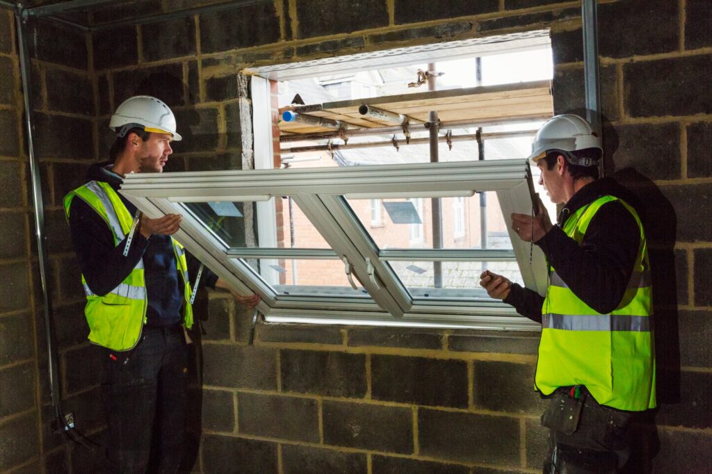Two builders in hard hats and high visibility jacket fitting a window frame.